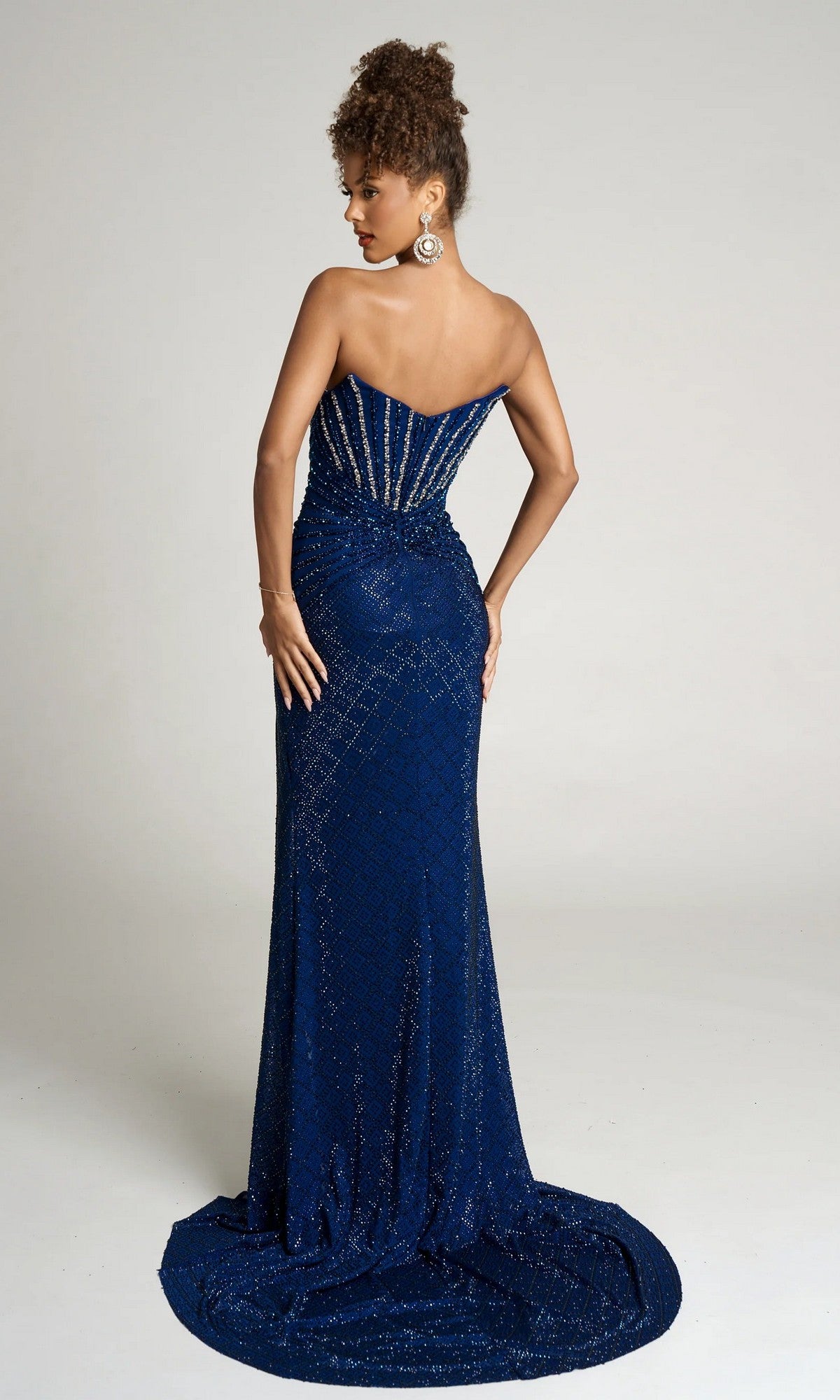 A woman models the Portia and Scarlett PS26016, a strapless, floor-length navy blue beaded gown with sparkling details, posing with her back to the camera against a plain background.